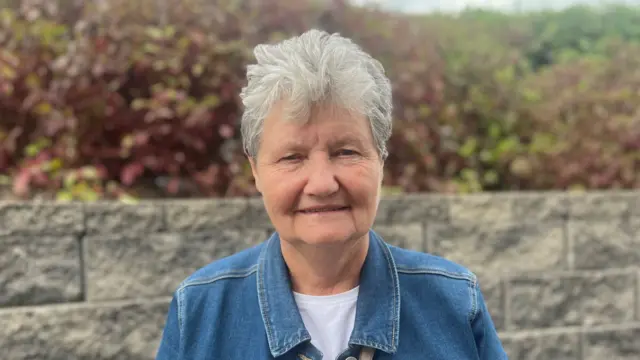 Margret in a denim jacket in front of a grey brick wall