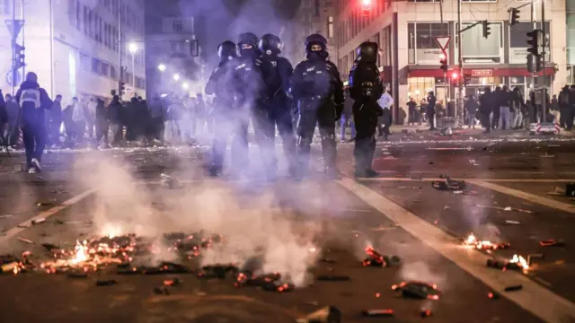Riot police stand guard on a street during New Year's Eve celebrations in Berlin. The ground is smoking as bits of detritus is still visibly alit.