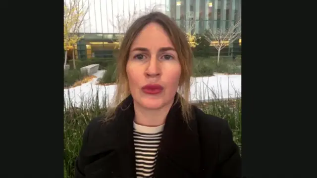 Missy Wilkinson appears wearing red lipstick, a black and white striped top and black coat. She stands in front of a building and what appears to be a path surrounded by greenery