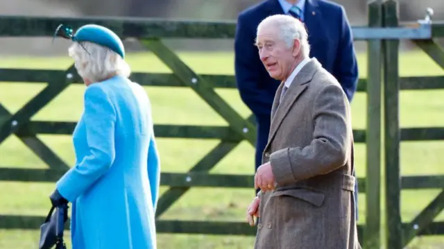 King Charles walking in countryside with Queen Camilla, both wearing long coats and holding large umbrellas