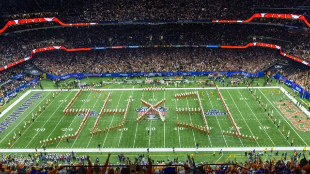 The Texas Longhorns marching band performing on the green pitch at the Caesars Superdome in New Orleans, with thousands in the stands