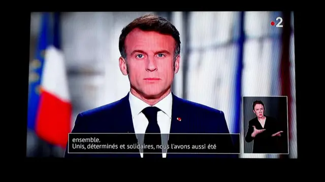 French President Emmanuel Macron is seen on a television screen as he delivers his New Year's speech, wearing a black suit and tie with white shirt in front of French and EU flags.