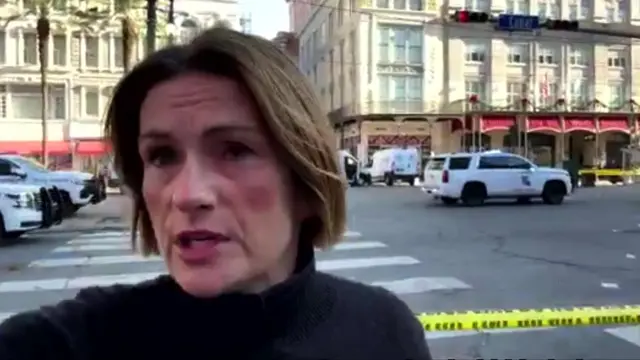 BBC reporter wears black turtleneck standing in front of street full of emergency vehicles and yellow police tape.