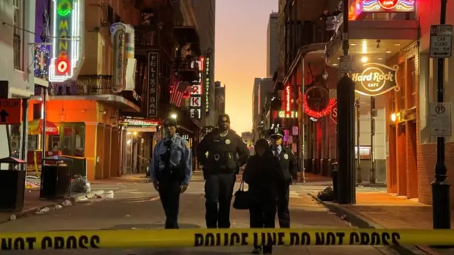Four police walk through the French Quarter, with LED signs on shop fronts, as the sun rises behind them