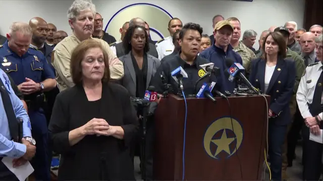 LaToya Cantrell speaking at a news conference with officials surrounding her and four microphones on a lectern in front of her