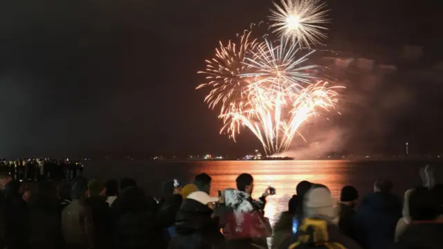 People can be seen taking pictures of the fireworks over a lake