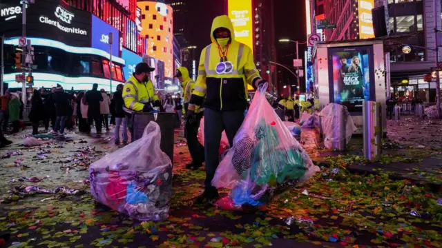 A sanitation worker dressed in yellow and carrying two bags of rubbish stands amid a sea of confetti in Times Square.