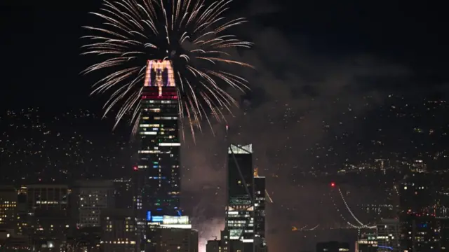 A firework display is seen over a darkened city skyline, with large skyscrapers in the foreground.