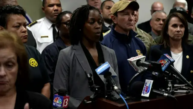 Alethea Duncan speaking at a news conference with officials stood around her, she is speaking into four microphones from different media