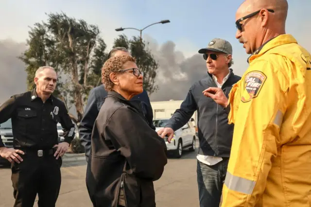 LA Mayor Karen Bass (and California Governor Gavin Newsom second on right) talk to a man wearing a bright yellow CalFire jacket. Smoke in background