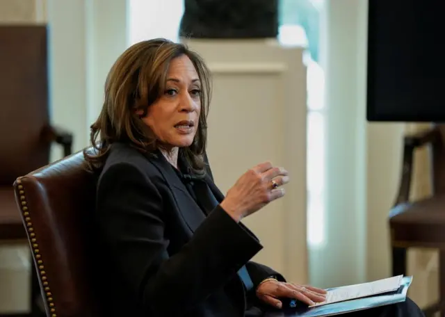 Kamala Harris sitting in a chair, balancing a notepad on her lap, as she speaks and holds one hand up