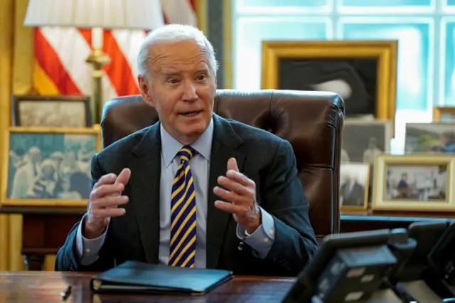 Biden sitting at a desk talking and gesturing with both hands. Lot's of family photos are behind him