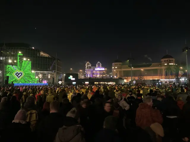 Thousands of people in an outside square wrapped up warm against the cold with bright light displays around them