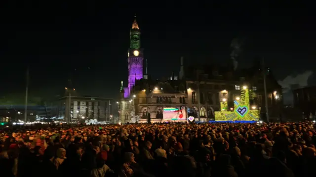 Thousands of people in a dark square with light installations look towards a stage