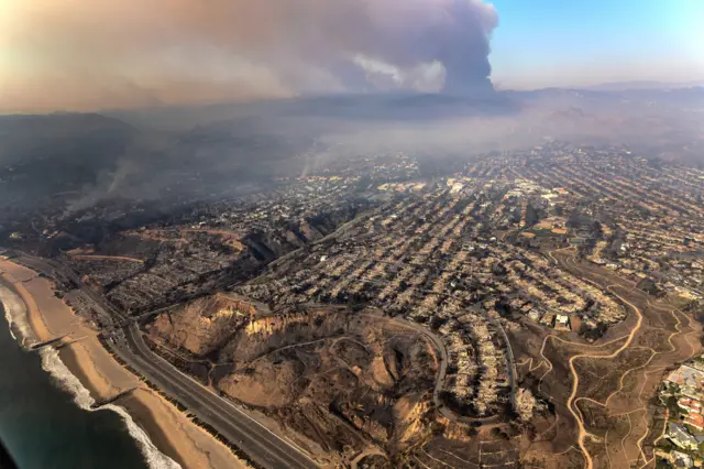 An aerial view shows a wide view of the Pacific Palisades neighbourhood, with rows of houses destroyed by the fires, and thick smoke in the hills in the background as fires continue.