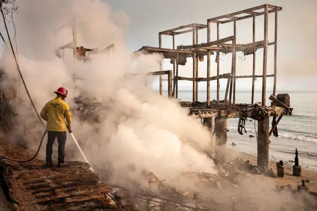 A firefighter points a water hose down at a smokldering section of a completely burnt out building by the sea in Malibu on Thursday