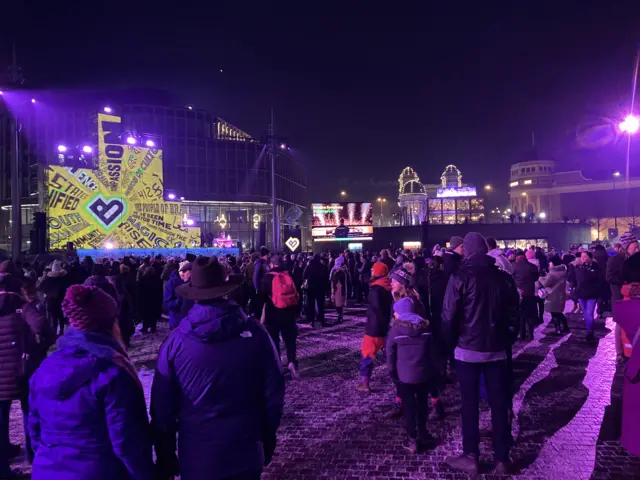 Thousands of people in an outside square wrapped up warm against the cold with bright light displays around them