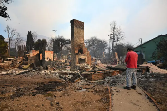 A man in a red top looks at the charred remains of a home, with the chimney breast and foundation the only remaining structures