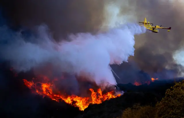 A yellow firefighting plane dumps a load of water over a fire burning in a valley