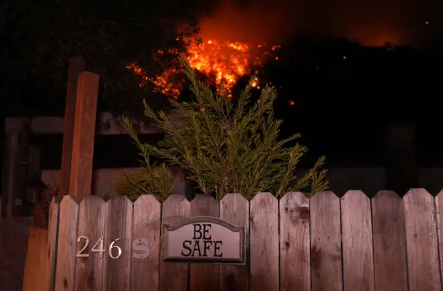 A wooden picket fence with teh sign 'be safe' and the Palisades fire clearly visible in the background