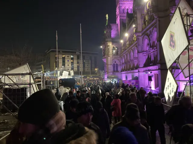 Thousands of people in an outside square wrapped up warm against the cold with bright light displays around them