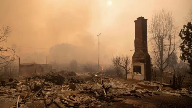 A building destroyed by fire with rubble on the ground, a chimney-like structure remains, haze and smoke covers the scene