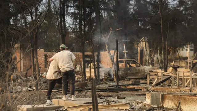 A man and a woman hug in front of a burnt-out building