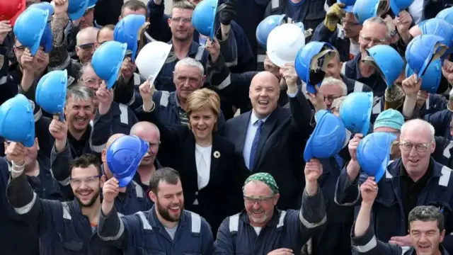 Nicola  Sturgeon and Jim McColl surrounded by shipyard workers raising their helmets in celebration