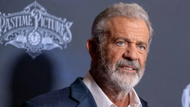 Mel Gibson in a shirt and jacket with an event backdrop behind