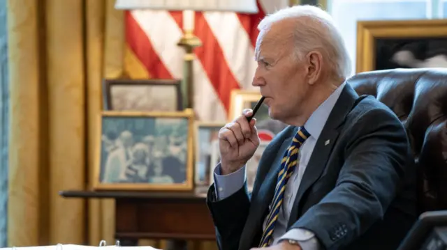 Joe Biden listens to video briefing sitting down at his desk inside the Oval Office. He's holding a pen to his lips while wearing a dark blue suit, striped blue and yellow tie and a shirt. Behind him, on a table in front of a window and yellow brocade curtains, are several family photos