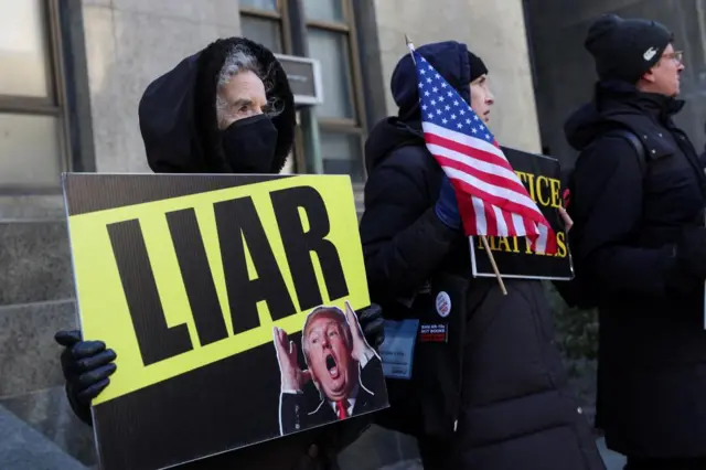 A woman in a jacket and face mask holds a sign saying liar with a picture of Trump. Someone stands next to her holding a flag that is covering the sign they hold