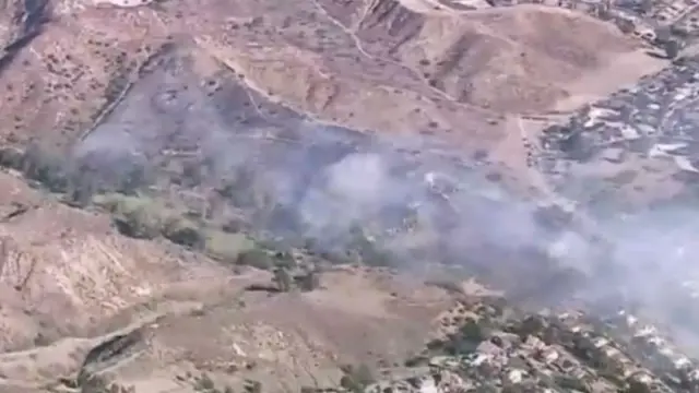 Fire and smoke in a brown hillside, houses in the bottom right corner