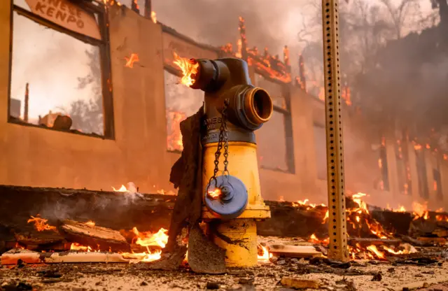 A fire hydrant on fire with one remaining wall of a building in the background on fire and debris alight on the ground