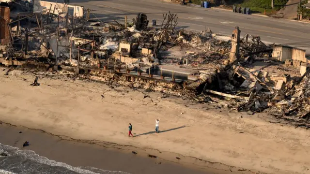 Aerial view of a row of burnt beachfront houses, with two people standing in front of them