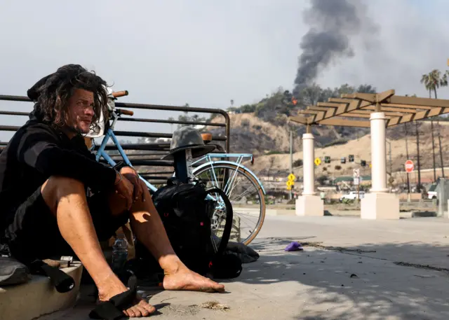 A man sits on the side of a road next to a blue bicycle as a fire burns in the hills behind him, in LA on Thursday