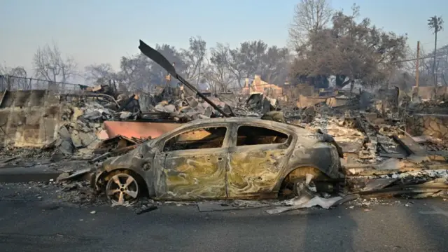 A burned out car stands next to a scene of rubble and ash in Altadena.