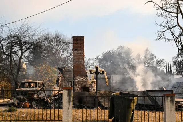 A chimney stands beside a burnt out car ot the left, and smouldering ruins to the right