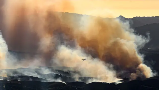 The silhouette of a plane is dwarfed by huge plumes of brown smoke rising out of a valley against a yellow sky