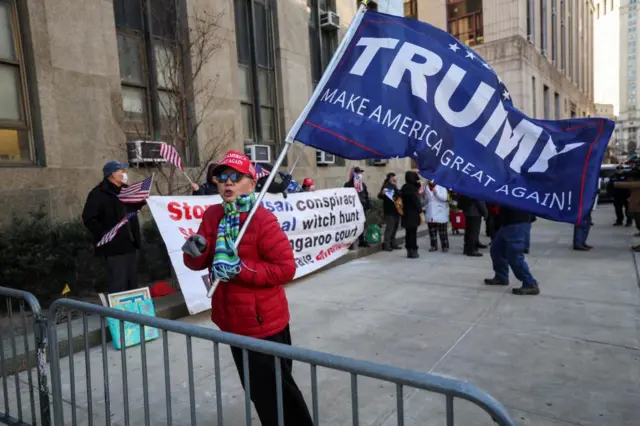 A woman by a fence holding a flag that says Trump on it, and behind her more people stand with banners and flags