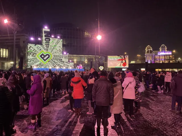 Thousands of people in an outside square wrapped up warm against the cold with bright light displays around them