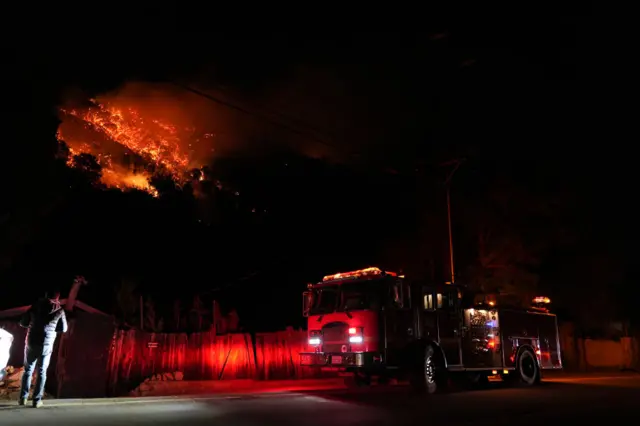 A fire truck stands next to a fence behind which a fiery blaze engulfs woodland. The image is shrouded in black with right lights blaring from the truck and flames.