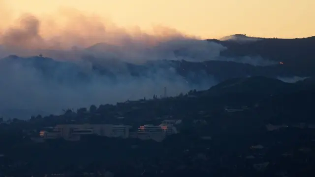 An aerial image shows fire and smoke behind the Getty Center Museum from wildfires, including the Palisades Fire, at sunset over Los Angeles, California, on January 8, 2025