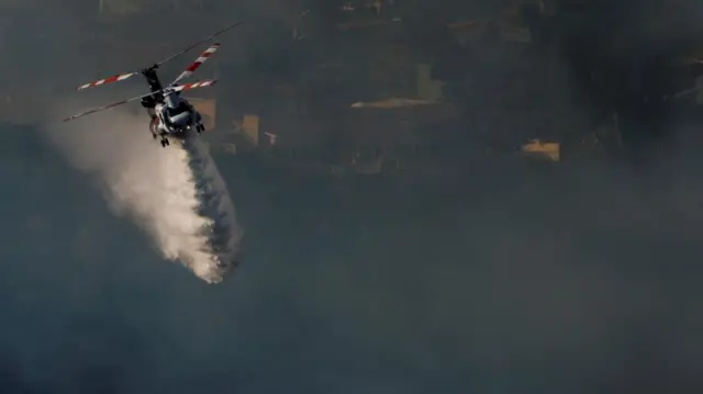 A helicopter dropping water on the ground below as thick smoke bellows in the air