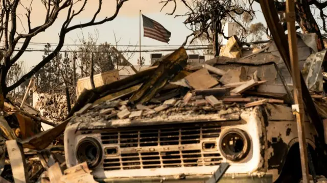 A burnt out truck covered in debris with a charred US flag flying on the background