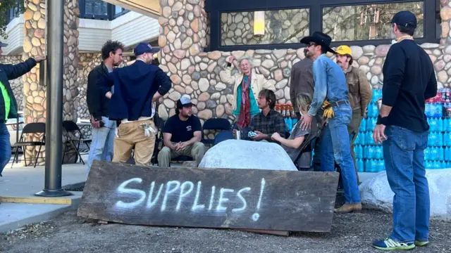 11 people stand and sit otuside a building, they have large stacks of bottle water behind them and a big, woooden, homemade sign reading "Supplies" spray painted on it