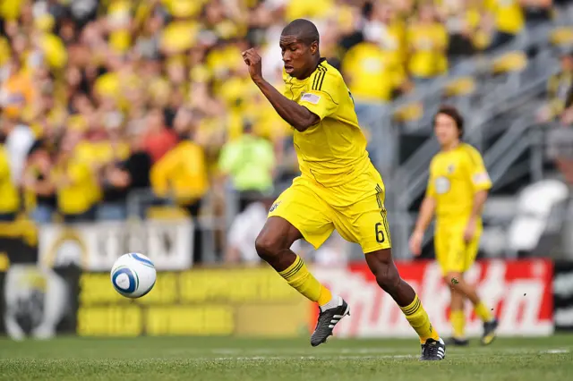 A footballer in a yellow kit with black boots chasing after a ball