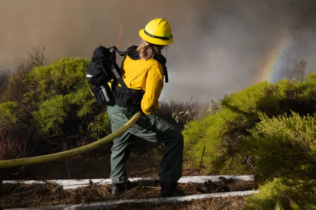 A firefighter works the Palisades Fire near the Sullivan Canyon area of Los Angeles, she is holding a hose, looking at smoke as a small rainbow is visible