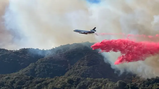 A firefighting aircraft flies over hills covered in trees and other plant life and is dumping a bright red fire retardant liquid below. Significant amounts of rising smoke is visible all around it.