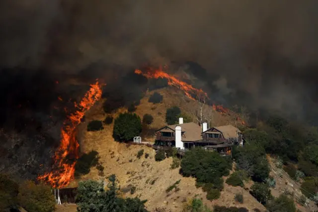 A hillside is on fire right above a large house, the flames are emitting so much smoke so little above can bee seen. The flames look to be encircling the property.