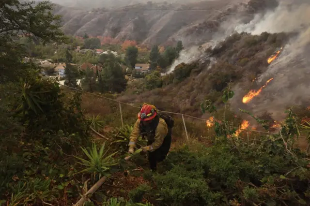 Firefighters deploy structure defense against the Palisades wildfire. A man is holding a line on a hillside and a fence is visible behind him.